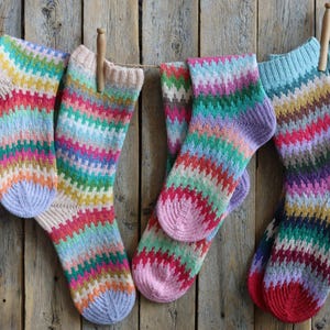 Pode incluir: Vários pares de meias coloridas e estampadas penduradas num varal contra um fundo de madeira rústica. As meias apresentam uma variedade de cores vibrantes, incluindo rosa, azul, verde e amarelo, criando uma estética alegre e acolhedora. Cada par é preso com um pregador de roupa de madeira.