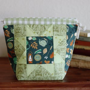 Può includere: Una pochette trapuntata con coulisse caratterizzata da un motivo centrale a stella. Il tessuto presenta uno sfondo verde scuro con illustrazioni di attrezzatura da campeggio, foglie e volti di animali. Il bordo superiore ha una finitura a quadretti.