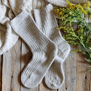 May include: A pair of light gray ribbed knit socks, resting on a rustic wooden surface. A cream-colored knitted blanket and yellow flowers with green stems are also present, creating a cozy, natural aesthetic.
