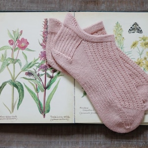 May include: A pair of light pink ankle socks with a textured knit pattern, resting on an open book with botanical illustrations. The socks are made of a soft material and are a muted rose color.