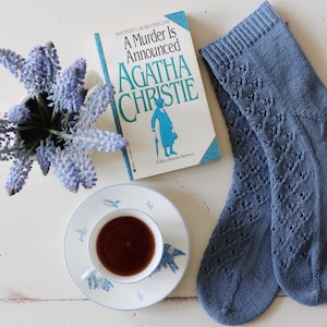 Puede incluir: Naturaleza muerta con un libro de Agatha Christie, una taza de té, un jarrón con flores azules y un par de calcetines azules de punto con un patrón decorativo. Los elementos están dispuestos sobre una superficie blanca.