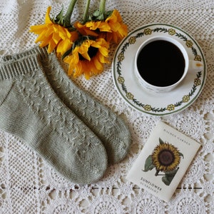 Op de afbeelding: Een paar groene gebreide enkelsokken met een delicaat patroon, een kop koffie op een schotel met een bloemenmotief en een zakje zonnebloempitten met de tekst "Halden Garden Sunflower Sunbright".