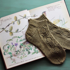 Könnte beinhalten: Ein Paar grüne gestrickte Kniestrümpfe mit Zopfmuster, die auf einem geöffneten Buch mit einer botanischen Illustration von Weidenzweigen und dem Wort "März" liegen.
