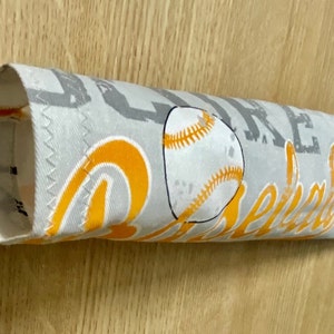 May include: A gray and white fabric baseball bat sleeve with orange and brown baseball and football graphics. The sleeve has the text "Game Day" printed on it.