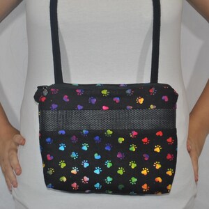 May include: A black shoulder bag with a rainbow paw print pattern. The bag has a mesh panel and a zipper closure.