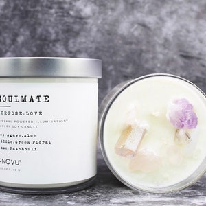 May include: A soy candle in a glass jar with a silver lid, featuring a white label with black text reading SOULMATE, PURPOSE.LOVE, MINERAL POWERED ILLUMINATION, LUXURY SOY CANDLE, 8.1 OZ | 240 G. The candle top contains embedded raw crystals.