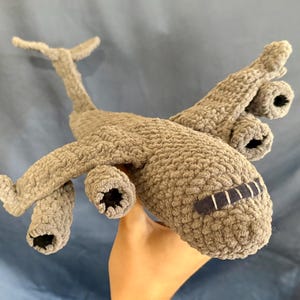 May include: A handmade crochet airplane toy crafted from textured light brown yarn. The plush aircraft features four distinct engine pods attached to the wings, a Y-shaped tail section, and a dark rectangular cockpit window with white stitched details.