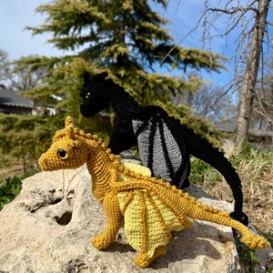 May include: Two crocheted dragon figures, one in mustard yellow and one in black with grey wing accents. Each dragon features a textured yarn body, pointed ears, a long tail, and large, round, dark eyes, standing in a side-profile pose.