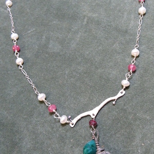 Puede incluir: Un collar de cadena de plata con cuentas blancas y rosadas y un colgante con una piedra verde y un diseño de jaula de plata.