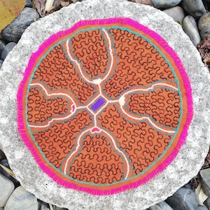 Könnte beinhalten: Ein runder, brauner Stoff-Patch mit einem geometrischen Muster, das in schwarzem, weißem und türkisfarbenem Garn gestickt ist. Der Patch ist von einem rosa und türkisfarbenen Rand umgeben und an einem grauen Stein befestigt.