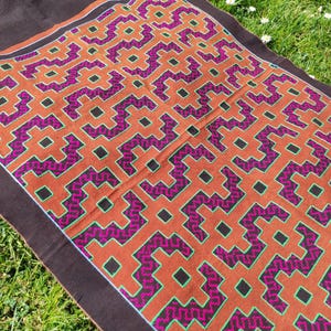 May include: A brown fabric tapestry with a geometric pattern in shades of orange, green, and purple. The pattern is made up of squares and rectangles, and it is repeated throughout the tapestry.