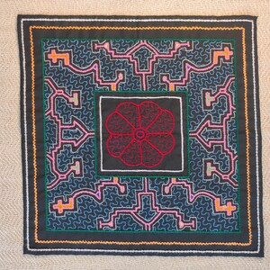 Op de afbeelding: Vierkante textiel met een zwarte rand en ingewikkelde geometrische patronen in roze, oranje en blauw op een donkerblauwe achtergrond. Een centraal vierkant heeft een rood bloemmotief. Het textiel is waarschijnlijk handgemaakt.
