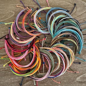 May include: A collection of multiple circular string bracelets arranged in a circular pattern. Each bracelet features a unique combination of vibrant colors including shades of blue, pink, orange, yellow, green, purple, and black, with knotted ends.