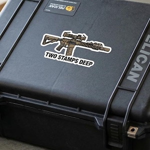 May include: A black Pelican case with a sticker that reads "TWO STAMPS DEEP" and a graphic of a rifle. A tan and black bag and a pair of olive green headphones are also visible. The case is open, revealing its interior.
