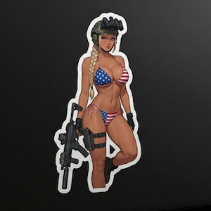 May include: A sticker featuring a cartoon illustration of a person in a bikini with an American flag design, holding a rifle. The sticker is on a black laptop, with an American flag in the background.