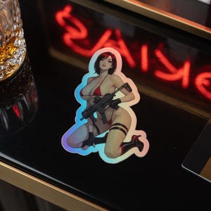 May include: A holographic sticker featuring an anime-style character with short dark hair, wearing a red bikini and black gloves, kneeling while holding a black submachine gun. The character has black thigh-high boots and a black strap on one leg.