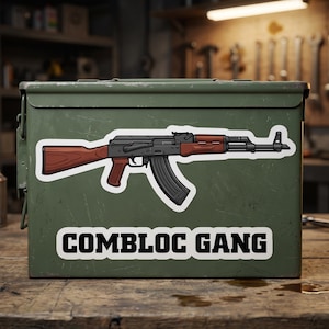 May include: An olive green metal ammunition box featuring a large sticker of a black and brown rifle with a curved magazine. Below the rifle, a white rectangular sticker displays the black text COMBLOC GANG in a bold, blocky font style.