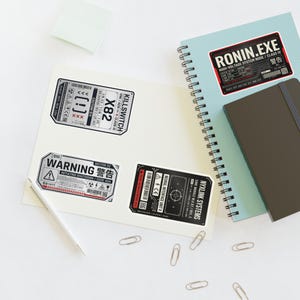 May include: A collection of tech-themed stickers with Japanese text and warning symbols. The stickers are black, white, and red, featuring text like "KILLSWITCH" and "WARNING". They are placed on a white surface with a pen, paperclips, and notebooks.