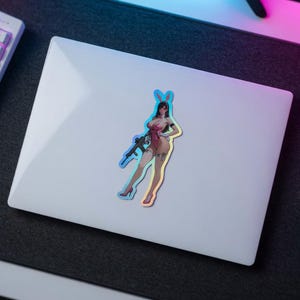 May include: A holographic sticker featuring an illustration of a character wearing a pink bunny suit, white gloves, and high heels, holding a black rifle. The character has long dark hair and bunny ears, with a visible tattoo on the left thigh.