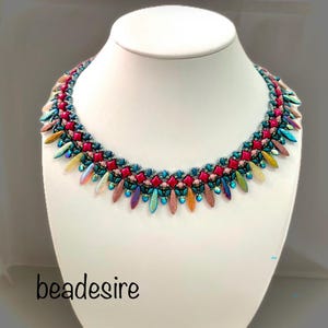 May include: A colourful beaded necklace with a striking design. The necklace features red, teal, and iridescent beads, with a teardrop-shaped fringe. The text "beadesire" is visible at the bottom.