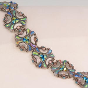 Puede incluir: Una pulsera de cuentas con un diseño floral repetido. Cada elemento presenta un grupo central de cuentas iridiscentes azules y verdes, rodeado de cuentas plateadas grises iridiscentes en forma de pétalo y pequeñas cuentas redondas. Fondo blanco.