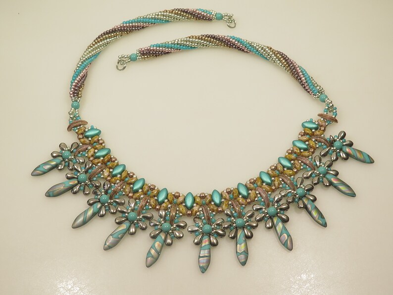 Peut inclure: Un collier de perles turquoise et or avec un motif g&eacute;om&eacute;trique. Le collier pr&eacute;sente une rang&eacute;e centrale de perles avec deux rang&eacute;es de perles plus petites de chaque c&ocirc;t&eacute;. Le collier est accentu&eacute; par des perles en forme de fleur turquoise et argent et de longues perles turquoise pointues.