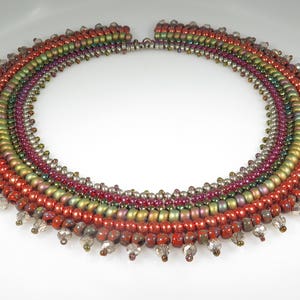 May include: A multi-strand beaded necklace with a graduated design. The necklace features a variety of colours, including red, green, and silver. The beads are arranged in a circular pattern, creating a unique and eye-catching design.
