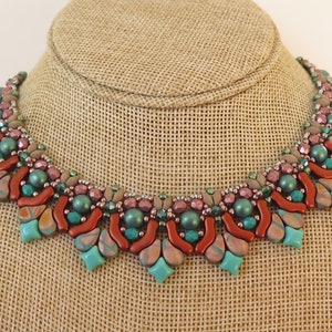 Puede incluir: Un collar de cuentas con un diseño geométrico en tonos turquesa, cobre y rosa. El collar se muestra en un maniquí de arpillera marrón.