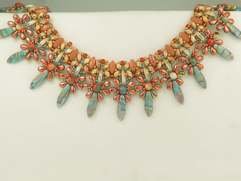 Peut inclure: Un collier de perles avec un design multi-brins pr&eacute;sentant un motif floral r&eacute;p&eacute;titif. Le collier est compos&eacute; d'un m&eacute;lange de perles brun clair, orange et bleues.