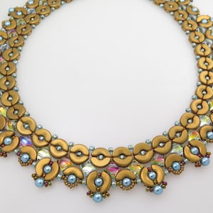 Puede incluir: Un collar de cuentas con un patrón circular de cuentas doradas mate en forma de media luna, cristales facetados transparentes iridiscentes y pequeñas cuentas redondas de color azul claro. El diseño está dispuesto en un estilo de collar simétrico.