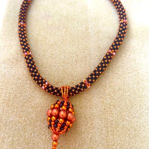 May include: A beaded necklace featuring a tubular band of small black and metallic orange beads. A central pendant hangs from the band, constructed from larger metallic orange and black beads in a rounded, textured, cage-like shape.