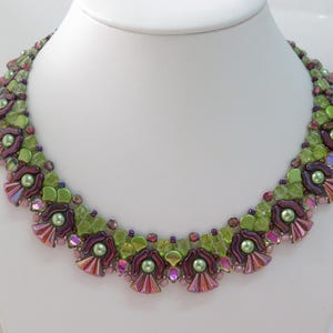 May include: A beaded necklace with a floral design. The necklace features a mix of green, purple, and iridescent beads. The design includes flower-shaped elements and small, round beads, creating a detailed and colorful accessory.