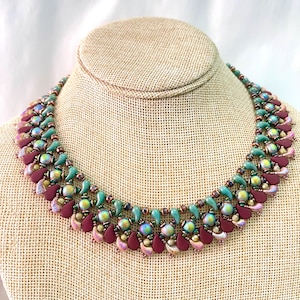 May include: A handcrafted necklace featuring a repeating pattern of burgundy, teal, and iridescent beads. The necklace is displayed on a neutral-toned bust, showcasing its intricate design and color palette. The necklace is a statement piece.