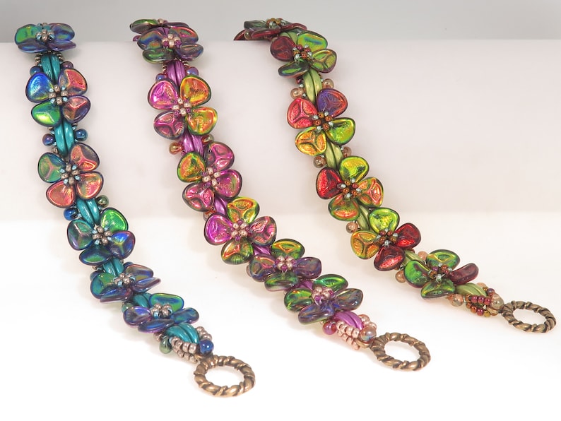 Peut inclure: Trois bracelets en perles avec un motif floral r&eacute;p&eacute;titif compos&eacute; de p&eacute;tales en verre iris&eacute; et multicolore. Chaque bracelet pr&eacute;sente un d&eacute;grad&eacute; de couleurs unique, allant du bleu et vert au violet, rose et dor&eacute;, termin&eacute; par un fermoir &agrave; bascule couleur bronze.
