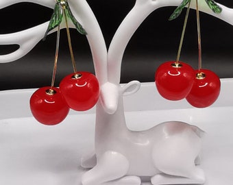 Peut inclure: Une paire de boucles d'oreilles en forme de cerises avec des feuilles vertes. Les boucles d'oreilles sont en résine rouge et dorée et sont suspendues à un support de boucles d'oreilles en forme de cerf blanc.