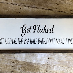May include: A white wooden sign with a brown frame, featuring the text "Get Naked" in a cursive font. Below the text, it reads "JUST KIDDING, THIS IS A HALF BATH. DON'T MAKE IT WEIRD."