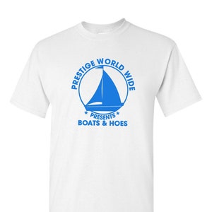 Op de afbeelding: Wit t-shirt met een blauwe afbeelding. De afbeelding toont een zeilboot in een cirkel met de woorden "PRESTIGE WORLD WIDE" eromheen. Onder de cirkel staat de tekst "PRESENTS BOATS & HOES".