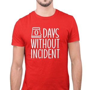 May include: A red t-shirt with white text and a graphic of a calendar with the number 0. The text reads "0 DAYS WITHOUT INCIDENT". The shirt is short-sleeved and appears to be made of a soft material.