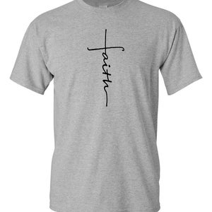 May include: A light gray t-shirt with a black cross design and the word "faith" written vertically. The shirt is made of a soft material and has short sleeves and a crew neck.