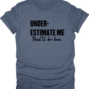 May include: A blue t-shirt with the text "UNDERESTIMATE ME That'll be fun." in black, centered on the front. The shirt has a crew neck and short sleeves, made of a soft, casual fabric.