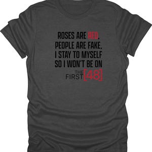 May include: Dark gray t-shirt with the text "ROSES ARE RED, PEOPLE ARE FAKE, I STAY TO MYSELF SO I WON'T BE ON THE FIRST 48" printed in black and red. The shirt is a crew neck style.