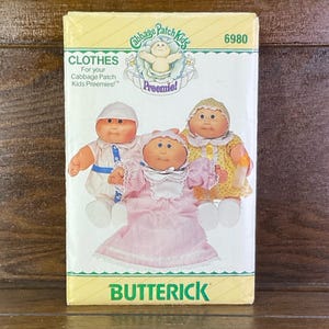 May include: A vintage sewing pattern for Cabbage Patch Kids Preemie dolls. The pattern is for making clothes for the dolls and features a picture of three Cabbage Patch Kids dolls wearing the outfits. The pattern is by Butterick and the number is 6980.