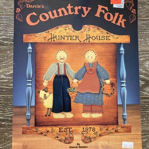 May include: A book cover for a painting pattern titled "Country Folk" featuring a wooden sign with the text "Hunter House" and the words "EST. 1978". The sign is painted with a blue and white design and features two figures, a sheep, and a wreath.