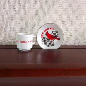 May include: A white ceramic mug and small plate set with a Great Smoky Mountains theme. The mug has "GREAT SMOKY MTS" in red lettering. The plate features a red cardinal bird and white flowers with gold trim.