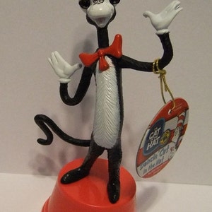 May include: A figurine of the Cat in the Hat character, standing on a red base. The figure is black with a white chest, white gloves, and a red bow tie. It wears a tall red and white striped hat. A tag reads "Dancin' Cat in the Hat".