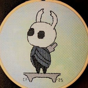 May include: A cross-stitch embroidery of a character from the video game Hollow Knight. The character is white with black eyes and a dark blue cloak, set against a light blue and green background. The embroidery is framed in a wooden hoop.