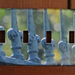 Könnte beinhalten: Eine dekorative Wandplatte mit einer Nahaufnahme eines blauen Metallzauns mit fünf dekorativen Endstücken. Der Zaun befindet sich vor einem verschwommenen grünen Hintergrund.