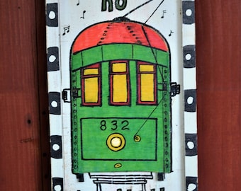 May include: A white wooden sign with black polka dots around the border. The sign has a green and red trolley painted on it with the words "It ain't no trolley dawlin'" written in black.