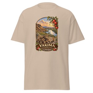 Peut inclure: T-shirt beige à manches courtes avec un motif représentant un paysage fluvial, des montagnes et un serpent. Le motif comprend le texte "YAKIMA WASHINGTON" dans une bannière. Des pommes rouges et des feuilles vertes ornent le haut du motif.