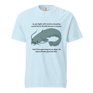 May include: Pale blue t-shirt with a gray catfish graphic and text. The text reads: "He who fights with monsters should be careful lest he thereby become a monster. And if thou gaze long into an abyss, the abyss will also gaze into thee."